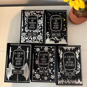 May include: A set of six black hardback books with silver foil detailing. The books are part of a fantasy series called "A Court of..." and feature titles like "A Court of Thorns and Roses", "A Court of Mist and Fury", "A Court of Wings and Ruin", "A Court of Frost and Starlight", and "A Court of Silver Flames".