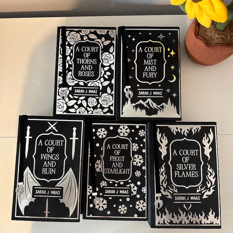 May include: A set of six black hardback books with silver foil detailing. The books are part of a fantasy series called "A Court of..." and feature titles like "A Court of Thorns and Roses", "A Court of Mist and Fury", "A Court of Wings and Ruin", "A Court of Frost and Starlight", and "A Court of Silver Flames".