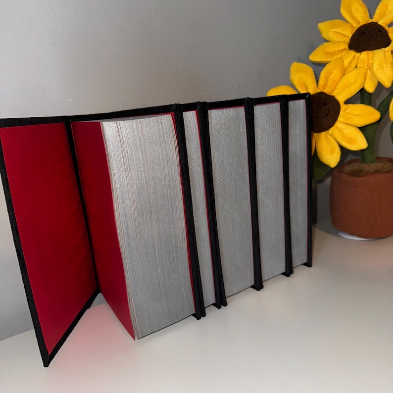 May include: A set of five books with black covers and red spines. The books are stacked on top of each other and are closed.