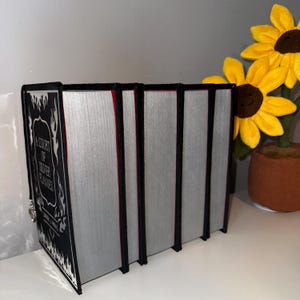 May include: A set of five hardback books with black covers and silver spines. The first book has a black and white illustration of flames and the text "A Court of Silver Flames".