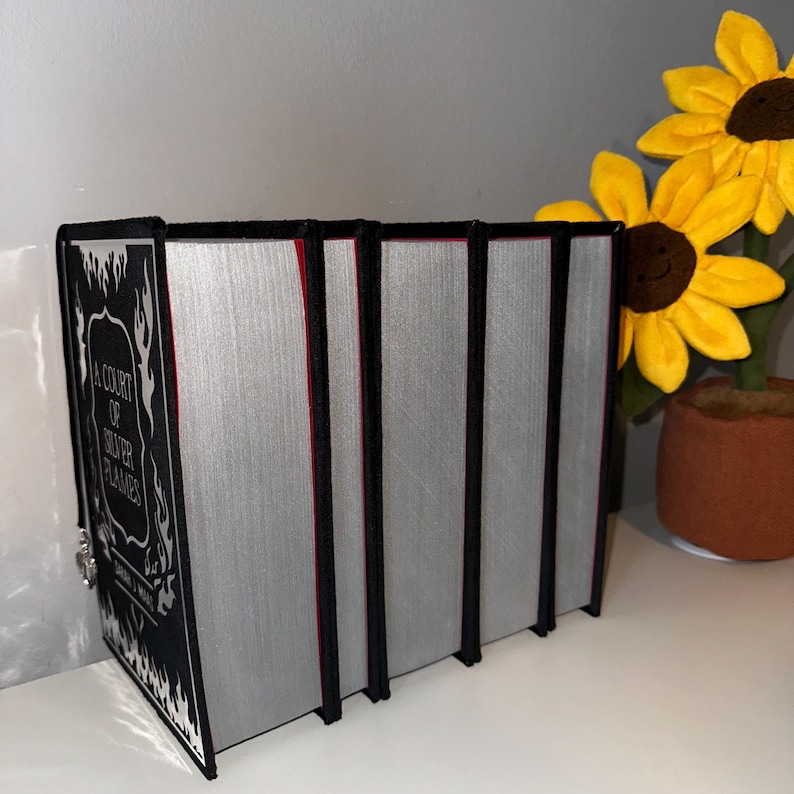 May include: A set of five hardback books with black covers and silver spines. The first book has a black and white illustration of flames and the text "A Court of Silver Flames".