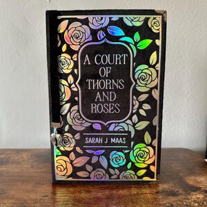 May include: A black hardback book with a holographic rose pattern and the title "A Court of Thorns and Roses" by Sarah J Maas.