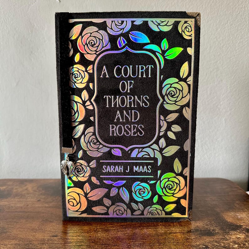 May include: A black hardback book with a holographic rose pattern and the title "A Court of Thorns and Roses" by Sarah J Maas.