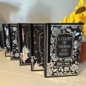 May include: A set of five black hardback books with silver foil accents and decorative bookmarks. The books are titled "A Court of Thorns and Roses" by Sarah J. Maas.