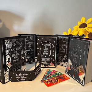 May include: A set of five black hardback books with silver foil detailing. The books are titled "A Court of Thorns and Roses", "A Court of Frost and Starlight", "A Court of Mist and Fury", "A Court of Silver Flames", and "A Court of Wings and Ruin". The books are arranged in a row on a white surface. There is a bookmark with the text "To the stars who listen" and a set of five bookmarks with the text "A Court of Thorns and Roses" in different colours.