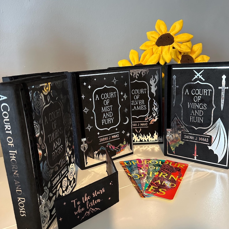 May include: A set of five black hardback books with silver and white designs. The books are titled "A Court of Thorns and Roses", "A Court of Mist and Fury", "A Court of Silver Flames", and "A Court of Wings and Ruin". There are also five colourful bookmarks with the same titles.