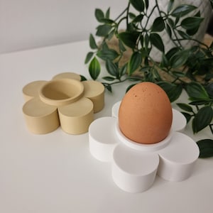 Könnte beinhalten: Zwei Keramik-Eierhalter in Form von Blumen. Einer ist weiß mit einem braunen Ei darin, der andere ist beige.