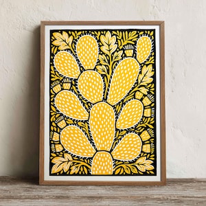 Puede incluir: Una lámina artística enmarcada que muestra un cactus de nopal estilizado en amarillo vibrante con detalles de puntos blancos. El cactus se sitúa sobre un fondo negro sólido, rodeado de intrincados patrones de hojas amarillas, en un marco de madera clara.
