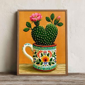 May include: A framed painting on an orange background featuring a green prickly pear cactus with a bright pink flower and buds, planted in a white ceramic mug decorated with colorful floral patterns in red, yellow, and green hues.