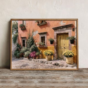 Op de afbeelding: Een ingelijste muurkunstprint met een gebouw in mediterrane stijl met perzikkleurige stucwerkmuren, een houten deur en een kasseien ondergrond. De scène bevat gele bloemen in potten, weelderige groene klimplanten en een zwarte wandlantaarn.
