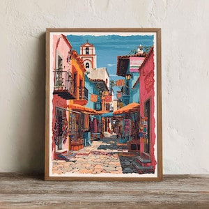 Könnte beinhalten: Ein gerahmter Druck einer farbenfrohen Straßenszene. Gebäude sind in Rosa-, Orange- und Blautönen bemalt, mit Markisen und Textilien. Ein Kirchturm ist im Hintergrund zu sehen, und die Kopfsteinpflasterstraße führt in die Ferne.