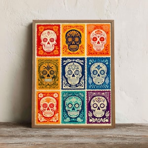 May include: A framed print featuring nine colorful Day of the Dead sugar skull designs. Each skull is intricately decorated with floral and geometric patterns, set against vibrant backgrounds of red, orange, yellow, blue, and purple.