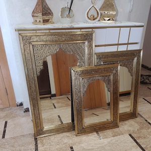 Op de afbeelding: Drie goudkleurige spiegels met gedetailleerde, sierlijke ontwerpen. De spiegels zijn rechthoekig met een gewelfde bovenkant en de frames hebben ingewikkelde patronen. Ze worden tentoongesteld op een lichtgekleurde tegelvloer.