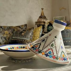 Peut inclure: Une tagine en céramique blanche avec un bord bleu et des motifs géométriques colorés dans des tons de rouge, orange, jaune, vert et bleu. La tagine est posée sur une assiette blanche avec un bord bleu.