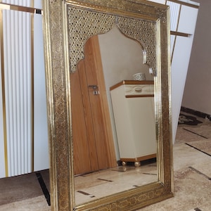 May include: An ornate gold-framed mirror with an arched top. The frame has detailed patterns. The mirror leans against a white cabinet with gold accents, reflecting a room with a wooden door and beige furniture.