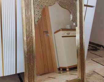 May include: An ornate gold-framed mirror with an arched top. The frame has detailed patterns. The mirror leans against a white cabinet with gold accents, reflecting a room with a wooden door and beige furniture.