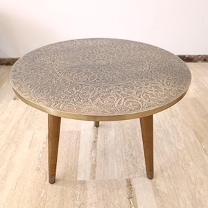 May include: A round coffee table featuring a metallic top with an intricate, engraved floral and geometric pattern in a bronze tone. The table is supported by three tapered wooden legs that have small, textured metallic accents at the base.