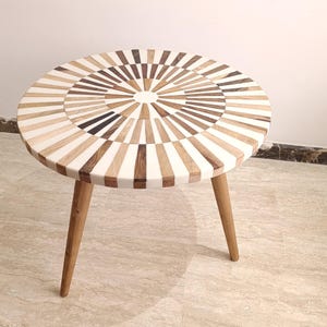 May include: A circular side table with a mosaic design on the tabletop. The top is patterned with wooden segments in brown, white, and black. It stands on three angled wooden legs. The table is about 50 cm in diameter.