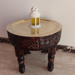 Pode incluir: Uma mesa lateral redonda de madeira castanha escura com padrões geométricos esculpidos e quatro pernas resistentes. A mesa apresenta um tampo de bandeja dourado com padrões. Uma lanterna branca com painéis de vidro amarelos está sobre a bandeja.