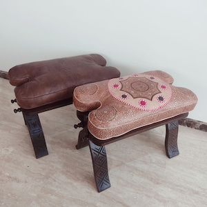 Peut inclure: Deux tabourets décoratifs en bois avec des dessus en cuir. L'un a un dessus en cuir marron, tandis que l'autre présente un dessus en cuir beige avec un motif complexe et des coutures roses. Les pieds en bois foncé ont des détails sculptés.