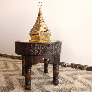 Peut inclure: Un petit tabouret rond en bois brun foncé avec des motifs géométriques sculptés sur le côté et quatre pieds tournés. Une lanterne en métal doré avec un dôme en forme d'oignon finement perforé repose sur la surface plane du tabouret.