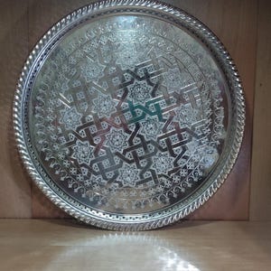 Può includere: Un vassoio rotondo in argento con un design intricato in rilievo. Il vassoio è rotondo e ha un bordo rialzato. Il design è un motivo ripetitivo di forme geometriche.