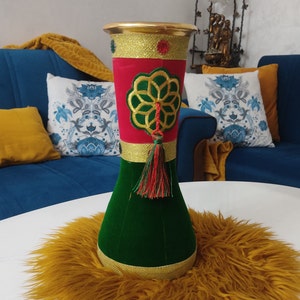 Peut inclure: Un vase décoratif vert, rouge et or avec un pompon. Le vase a un motif géométrique et repose sur une surface dorée et moelleuse.
