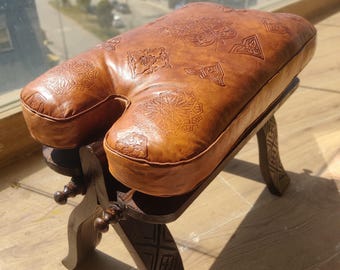 May include: A tan leather stool with a U-shaped seat featuring embossed geometric patterns. The seat rests on a dark brown wooden base with four carved legs that have intricate linear designs and small decorative knobs on the frame.