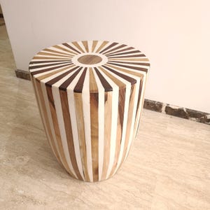 Peut inclure: Un tabouret décoratif en bois avec un motif en forme d'éventail sur le dessus. Le dessus présente des rayures alternées marron foncé, marron clair et blanches rayonnant d'un cercle central. Les côtés ont des rayures verticales dans la même gamme de couleurs.