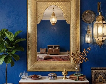 May include: A rectangular wall mirror with a wide, textured gold frame featuring an arched top. The mirror reflects a room with a wooden chest, patterned pillows, and a hanging lantern. It is mounted on a deep blue wall above a wooden console table.