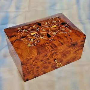 Peut inclure: Une boîte en bois avec un incrustation décorative de motifs en spirale et des accents dorés. La boîte a un trou de serrure et une clé en laiton.