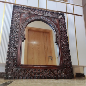 Op de afbeelding: Een rechthoekige, donkerbruine houten spiegel met een gewelfde bovenkant. Het frame heeft ingewikkelde, gesneden geometrische patronen en bloemmotieven. De spiegel reflecteert een houten deur en een interieur.