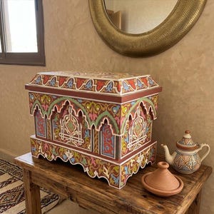 May include: A rectangular wooden chest featuring intricate hand-painted patterns in shades of red, green, blue, and yellow, with arched architectural motifs. Beside it sits a ceramic teapot and a small terracotta tagine lid on a wooden table.