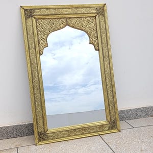 Peut inclure: Un miroir rectangulaire doré avec un sommet en forme d'arche. Le cadre présente des motifs et des dessins complexes et répétitifs. Le miroir reflète un ciel nuageux, ajoutant de la profondeur. Le miroir est appuyé contre un mur blanc.