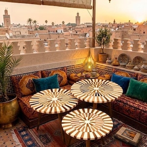 Peut inclure: Une terrasse sur le toit avec trois tables rondes en bois au motif incrusté en rayons de soleil, disposées sur un tapis à motifs. L'espace comprend des sièges intégrés avec des coussins orange et bleus, des lanternes et une vue urbaine.