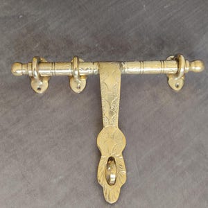 Peut inclure: Un loquet de porte doré avec un design orné. Le loquet a une longue barre horizontale avec deux boutons ronds à chaque extrémité. La partie verticale du loquet a un motif floral décoratif.