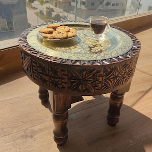 May include: A round, dark brown wooden side table with carved floral patterns. The table has a decorative brass tray on top, holding a plate of chocolate chip cookies and a glass teacup. The table is supported by four sturdy legs.