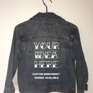 May include: A dark blue denim jacket hanging on a black hanger. The back features white text that reads YOUR IDEA HERE and CUSTOM EMBROIDERY ORDERS AVAILABLE. The jacket has a classic collar, long sleeves with button cuffs, and two chest pockets.