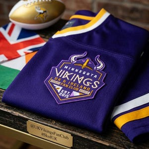 May include: A purple jersey with white and yellow sleeve stripes featuring a Minnesota Vikings UK & Ireland Fan Club patch with the text EST. 2015. A gold and white football with a Vikings logo and a flag are visible in the background.
