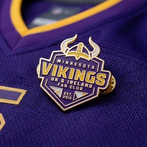 May include: A purple and gold metal pin featuring a Viking helmet icon and the text Minnesota Vikings UK & Ireland Fan Club Est. 2015, resting on a purple jersey with a gold-trimmed collar and a partial gold number 5 visible on the fabric.