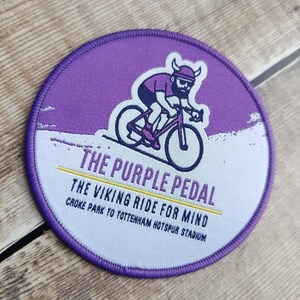 May include: Round purple patch with a cyclist graphic and the text "THE PURPLE PEDAL". The cyclist wears a horned helmet and rides a purple bicycle. The text "THE VIKING RIDE FOR MIND" and "CROKE PARK TO TOTTENHAM HOTSPUR STADIUM" are also included.