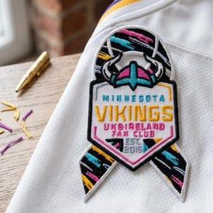 May include: A white jersey featuring an embroidered patch shaped like a ribbon. The patch displays a Viking helmet icon above the text MINNESOTA VIKINGS UKIRELAND FAN CLUB EST. 2015, rendered in yellow, pink, and light blue on a black background.