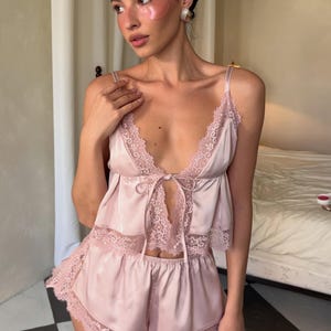 Puede incluir: Un conjunto de pijama de sat&eacute;n rosa p&aacute;lido compuesto por una camisola y pantalones cortos. La camisola presenta un escote en V profundo con delicados adornos de encaje, tirantes finos y un cierre de lazo frontal. El pantal&oacute;n corto tiene encaje.