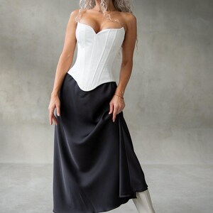 May include: A white corset top with a sweetheart neckline paired with a long, black satin skirt. The outfit is completed with white boots. The overall look is elegant and stylish, suitable for formal occasions.