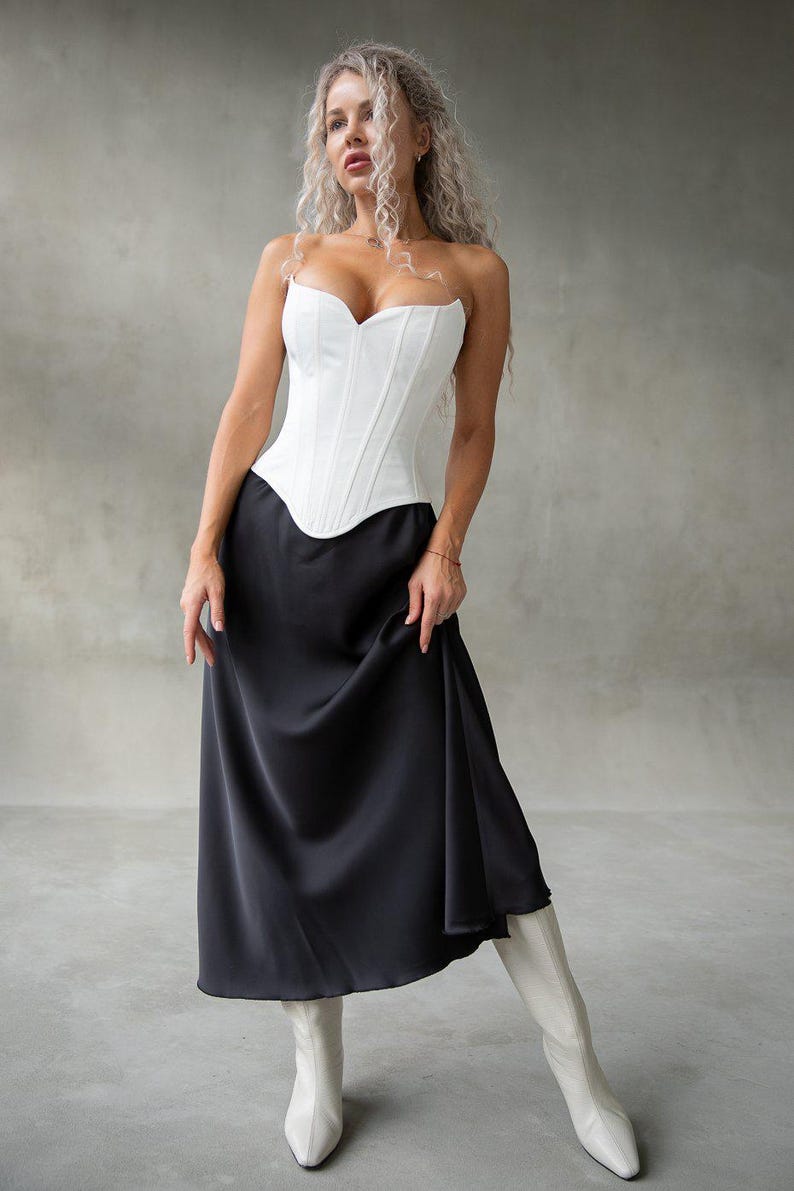 May include: A white corset top with a sweetheart neckline paired with a long, black satin skirt. The outfit is completed with white boots. The overall look is elegant and stylish, suitable for formal occasions.