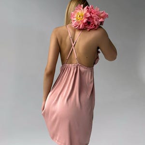 May include: A dusty rose-colored slip dress with spaghetti straps and a gathered neckline. The dress is made of a silky material. A bouquet of pink peonies rests on the back of the dress.