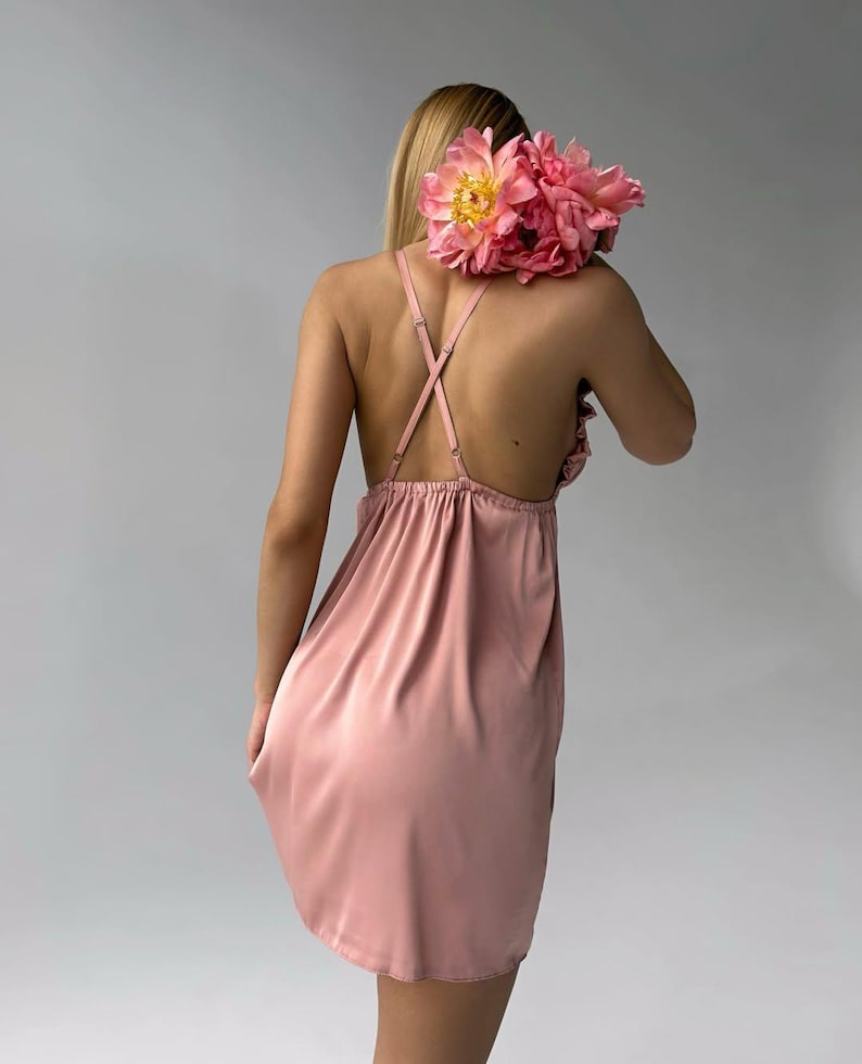 May include: A dusty rose-colored slip dress with spaghetti straps and a gathered neckline. The dress is made of a silky material. A bouquet of pink peonies rests on the back of the dress.