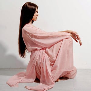 May include: A pale pink, flowing dress with long sleeves and a draped waist. The dress is made of a soft, lightweight fabric. The model is sitting on the floor with long, dark hair.