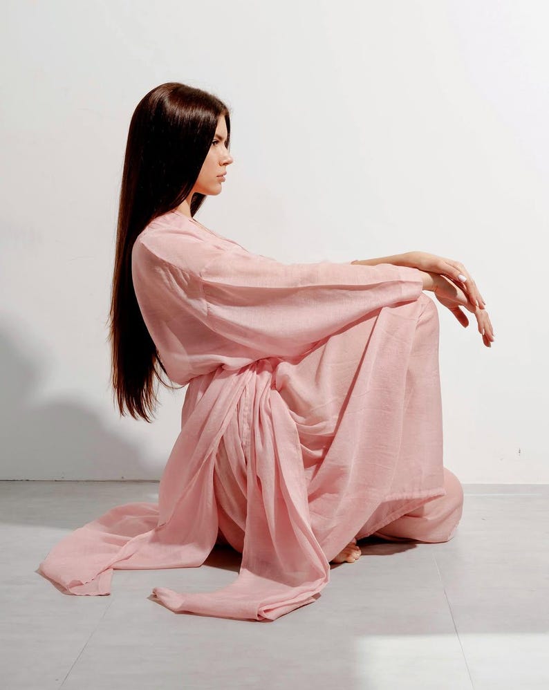 May include: A pale pink, flowing dress with long sleeves and a draped waist. The dress is made of a soft, lightweight fabric. The model is sitting on the floor with long, dark hair.