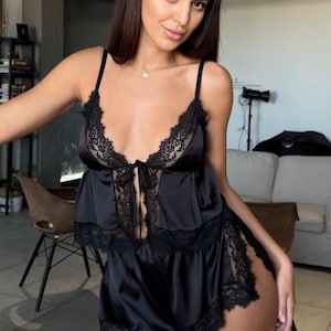 May include: A black two-piece pajama set consisting of a camisole top and matching shorts. The camisole features thin shoulder straps, a deep V-neckline, and black lace trim. The shorts have a high-cut leg design with matching black lace trim.